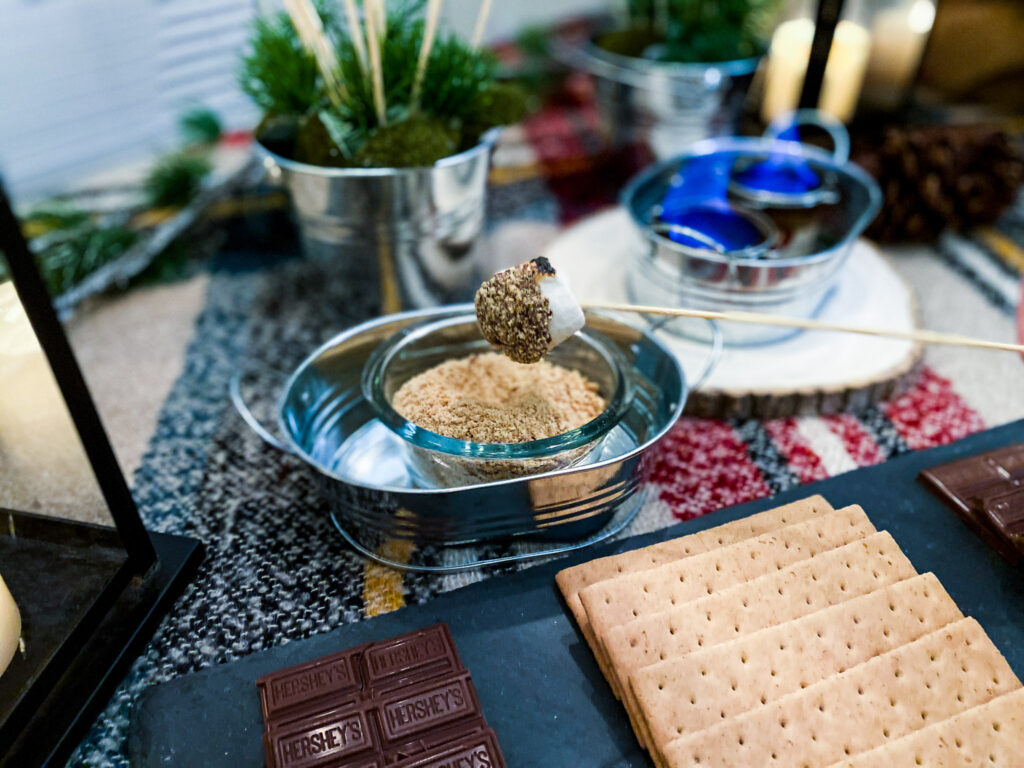 A toasted marshmallow is dipped in boozy chocolate fondue recipe, and then crushed graham cracker crumbs. It's a great s'mores dip recipe and a campfire party idea. It not only tastes fantastic, it looks cure in this galvanized pail on top of a plaid tablecloth and pinecone, with are great camping themed party decorations and camping themed party ideas. They also work great for a campfire party and are easy campfire party ideas. You could also use this as a campfire birthday party ideas.