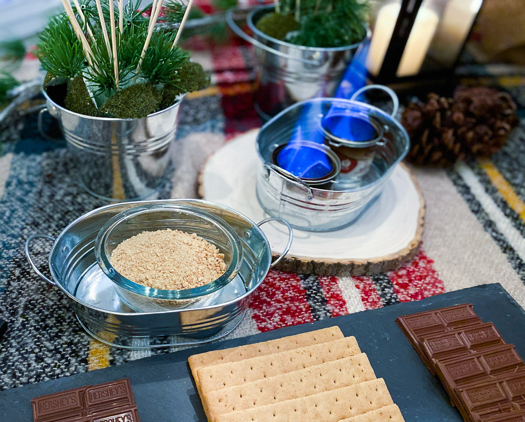 These adorable camping themed party ideas and camping themed party decorations include repurposing a plaid scarf as a tablecloth, black lanterns, marshmallow bouquets, galvanized serving pieces and a s'mores bar station. It's a great campfire party ideas with camping themed party food. S'more ingredients are displayed on a slate tray. S'mores dip is also included as one of the campfire party ideas. There is a lot of cuteness going on with this campfire birthday party ideas, great campfire party.