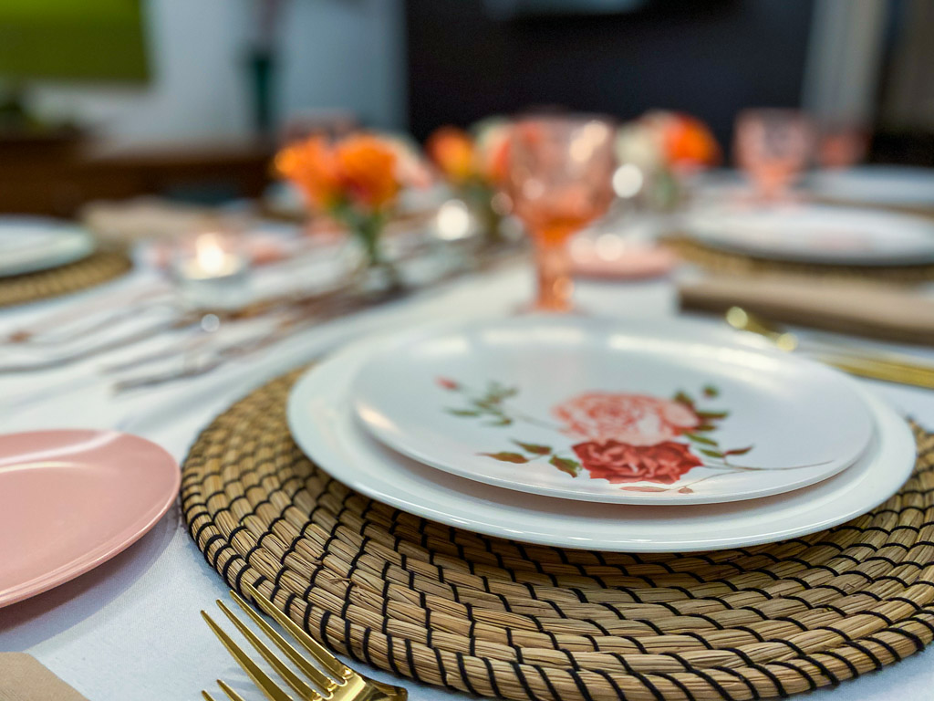 Colored glassware on a vintage tabletop with melamine pates and seagrass placemats, gold utensils and coral flowers.