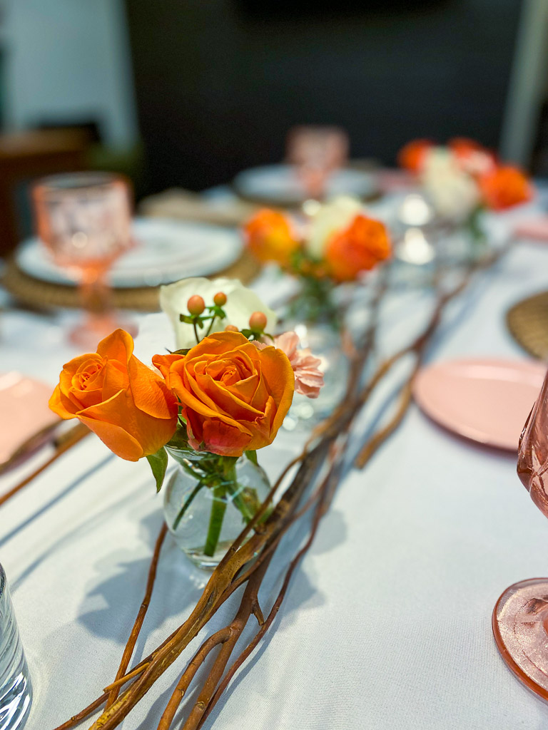 Coral roses in a small bud vase, surrounded by wooden twigs is the perfect complement to the vintage colored glassware on the tabletop.