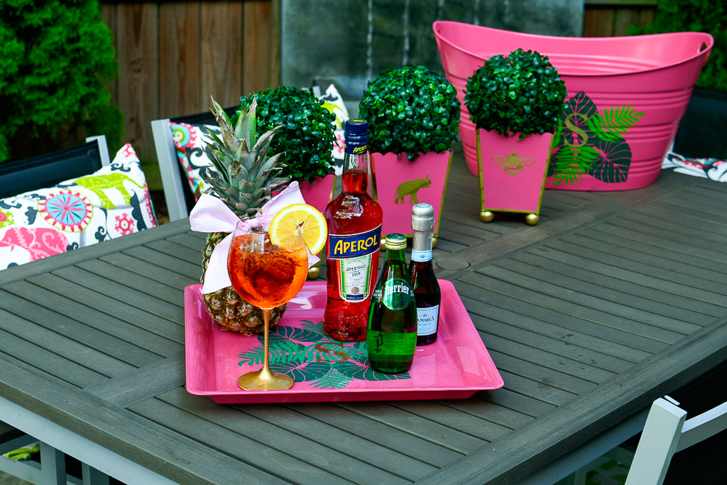 An Aperol Spritz cocktail with an Aperol bottle, sparkling Perrier water and a split of prosecco, pineapple with cute bow, hot pink tray with palm leaves and monogram, how to make an Aperol spritz, aperol spritz ratios, DIY cachepot, cute patio decor