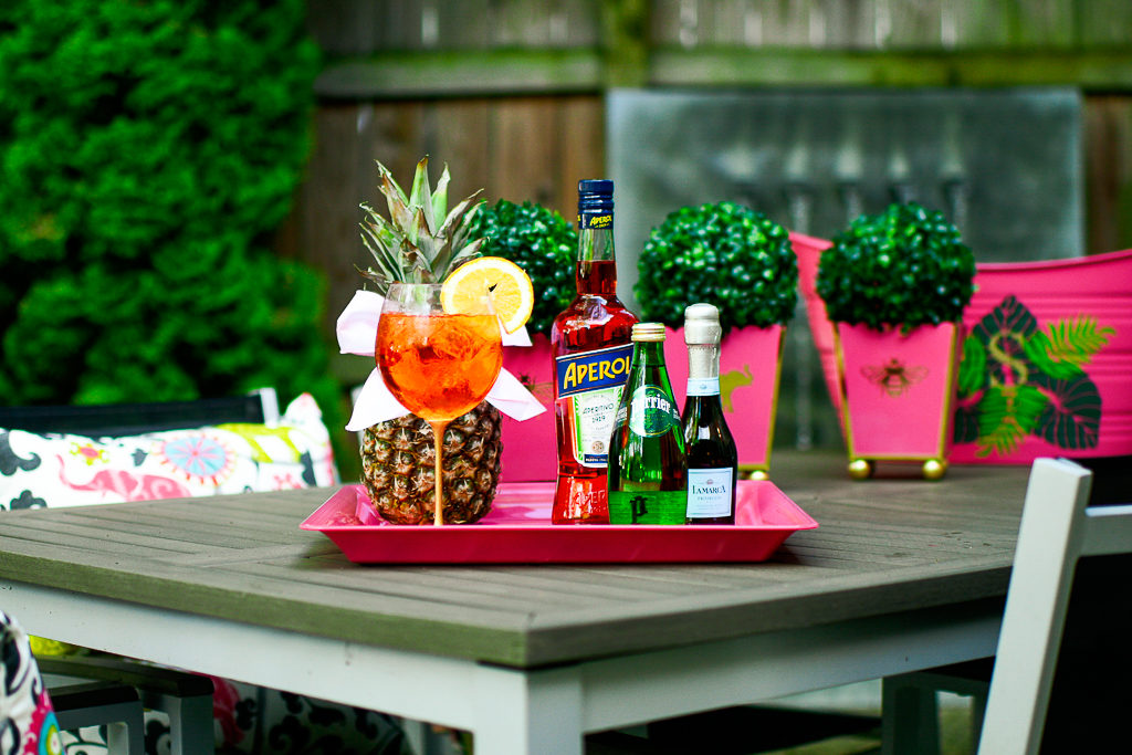 An Aperol Spritz cocktail with an Aperol bottle, sparkling Perrier water and a split of prosecco, pineapple with cute bow, hot pink tray with palm leaves and monogram, how to make an Aperol spritz, pink cachepots, boxwood, patio, aperol spritz ratios.