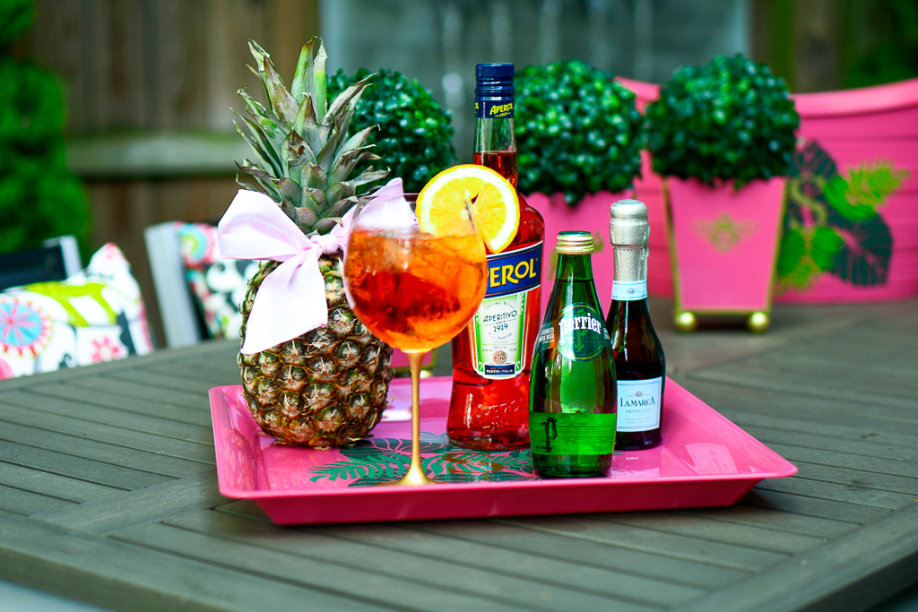 An Aperol Spritz cocktail with an Aperol bottle, sparkling Perrier water and a split of prosecco, pineapple with cute bow, hot pink tray with palm leaves and monogram, how to make an Aperol spritz