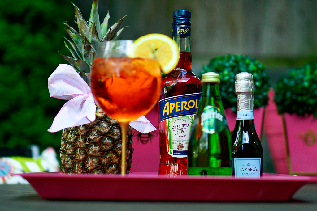 An Aperol Spritz cocktail with an Aperol bottle, sparkling Perrier water and a split of prosecco, pineapple with cute bow, hot pink tray with palm leaves and monogram, how to make an Aperol spritz, Aperol Spritz recipe, Aperol spritz