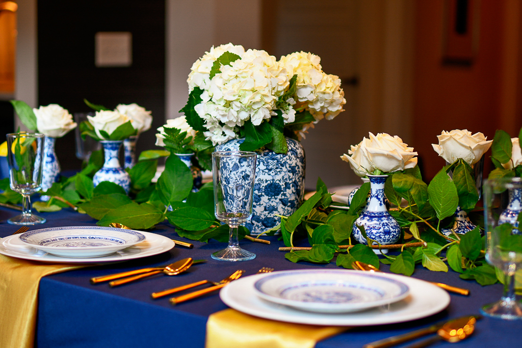 dinner table with navy tablecloth, chinoiserie ginger jar with white hydrangeas, chinoiserie bud vases with white roses, blue table decorations, blue table setting ideas 