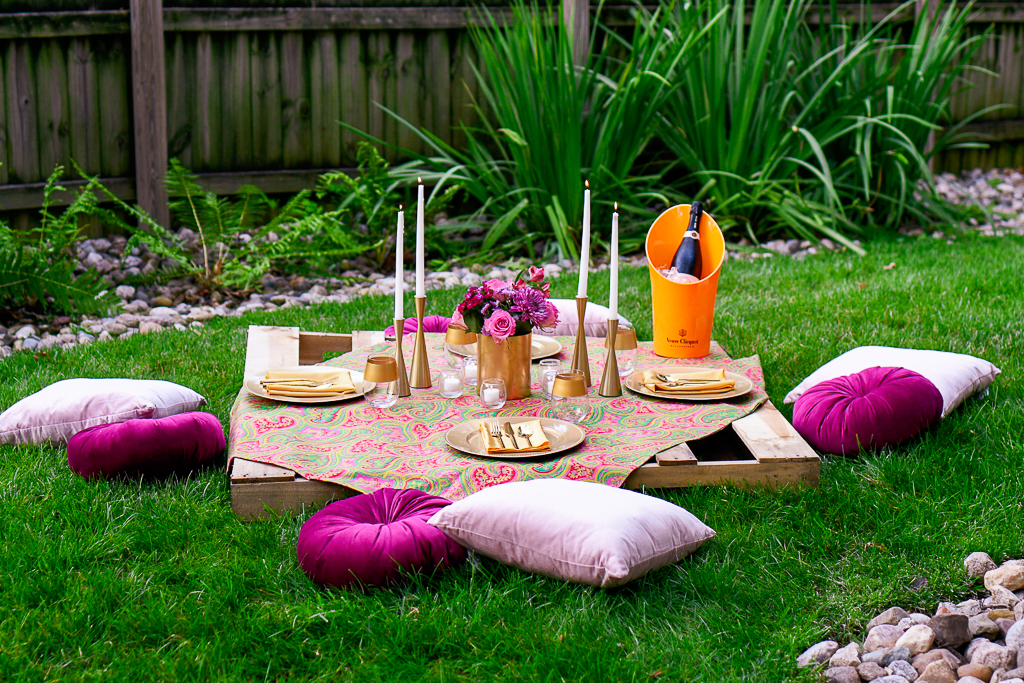 A picnic table that is a wooden pallet, with gold plates, napkins and gold utensils, pale pink velvet pillows, magenta velvet pillows, gold candlesticks, veuve clicquot, and white tapers. Romantic picnic ideas.