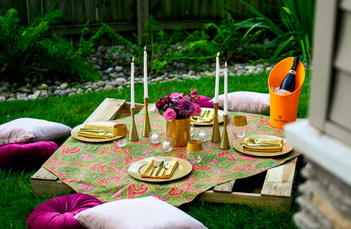 A pallet picnic table sits on green grass with gold chargers, gold flatware, gold candlesticks, white tapers, velvet cushions and a gorgeous bouquet of pink rose, purple daisy, and greenery in a gold vase. Romantic picnic ideas.