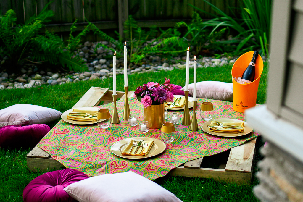 A pallet picnic table sits on green grass with gold chargers, gold flatware, gold candlesticks, white tapers, velvet cushions and a gorgeous bouquet of pink rose, purple daisy, and greenery in a gold vase. Romantic picnic ideas.