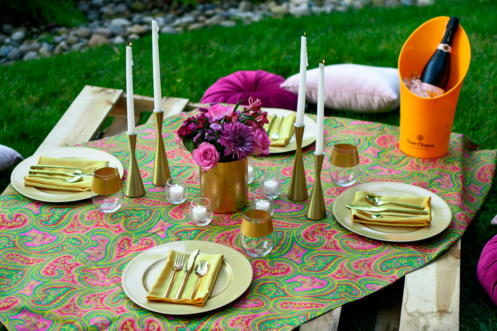 A picnic tablescape with a wooden pallet, gold chargers, gold napkins, gold utensils, white taper candles, gold candleholders, velvet pillows as cushions and gold rimmed wine glasses.