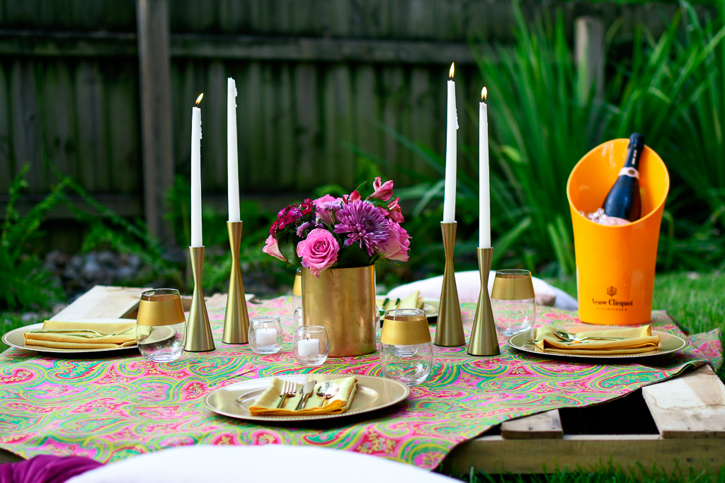 This simple picnic table setting uses a wooden pallet as a table and contains gold candlesticks, gold chargers, gold utensils, gold napkins and a fun paisley table linens. It's an inexpensive and romantic picnic ideas.