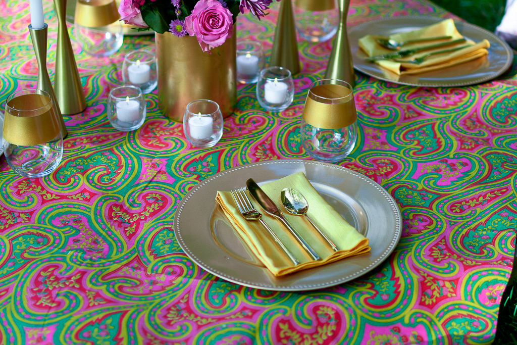 A gorgeous picnic tablescape with gold utensils, gold charger, gold rimmed wine glass and a fun paisley table linen. 