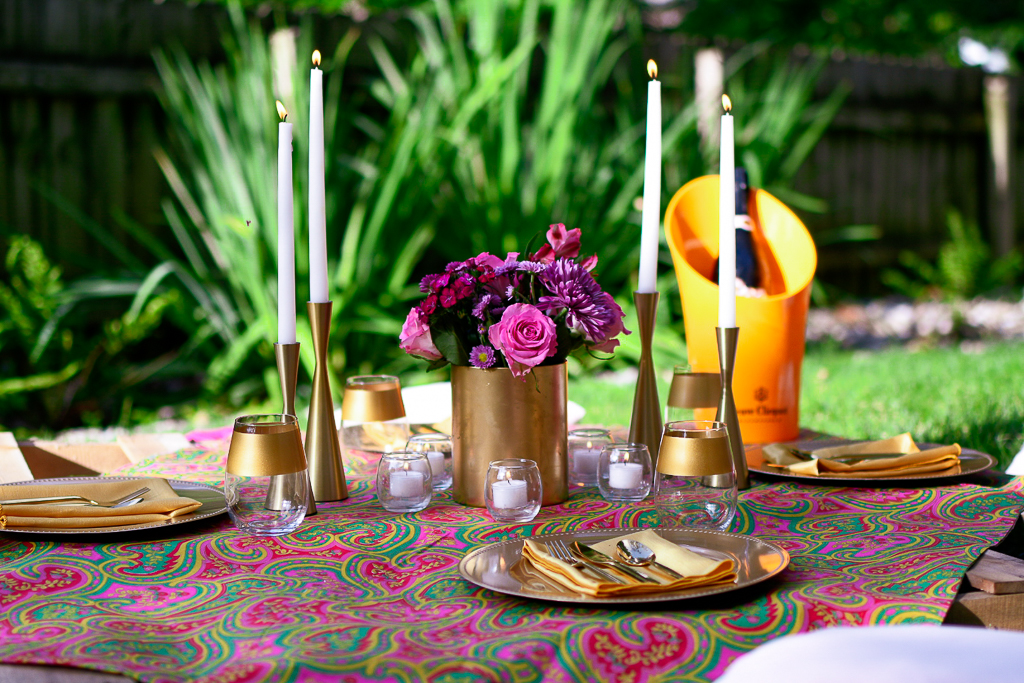 A gorgeous picnic tablescape with gold plates, gold napkins, gold utensils and gold rimmed wine glasses, as well as votive candles, gold candlesticks and white tapers. Easy picnic ideas for couples.